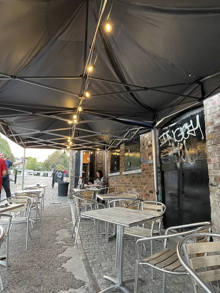 Outdoor tables and chairs under a black canopy strung with warm bulbs beside the canal towpath