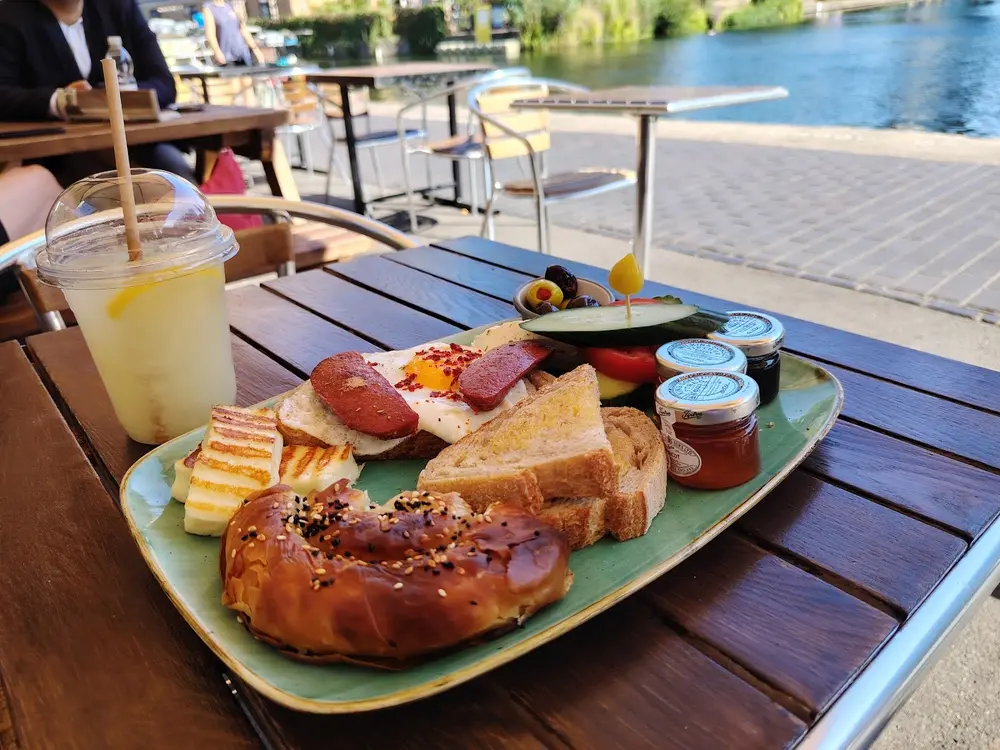 Turkish breakfast plate and a lemonade on a wooden table beside the canal, with the water and towpath behind